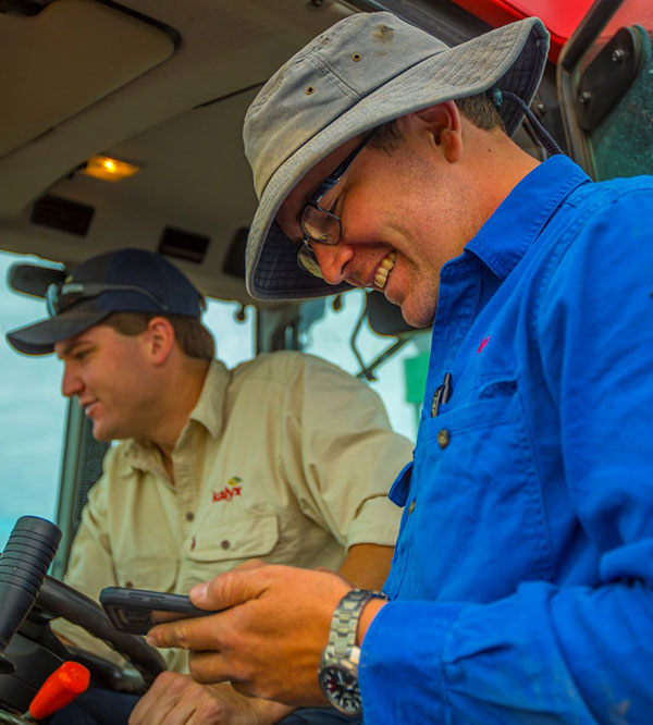 Two workers in a tractor cabin, one using a smartphone.